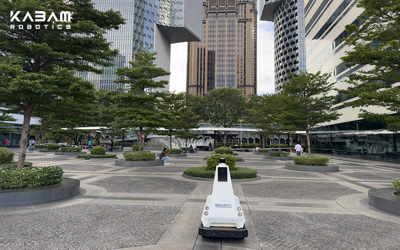 KABAM outdoor security robot patrolling a public plaza surrounded by modern buildings and greenery